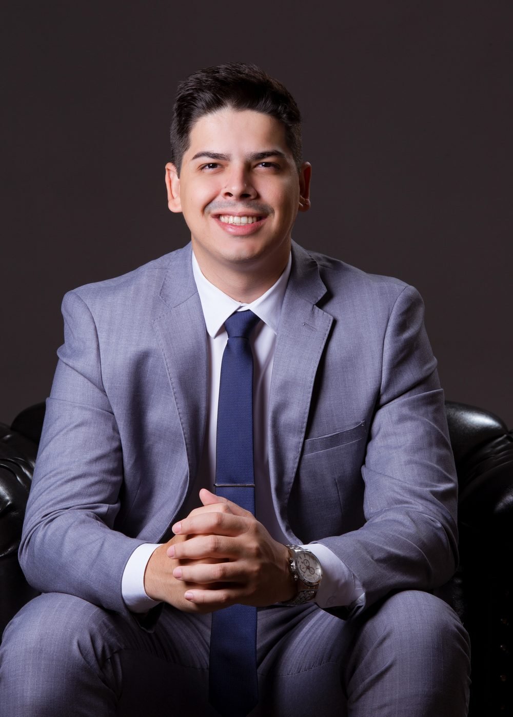 Dr. Mateus Moreira, de terno cinza e gravata azul, sentado e sorrindo